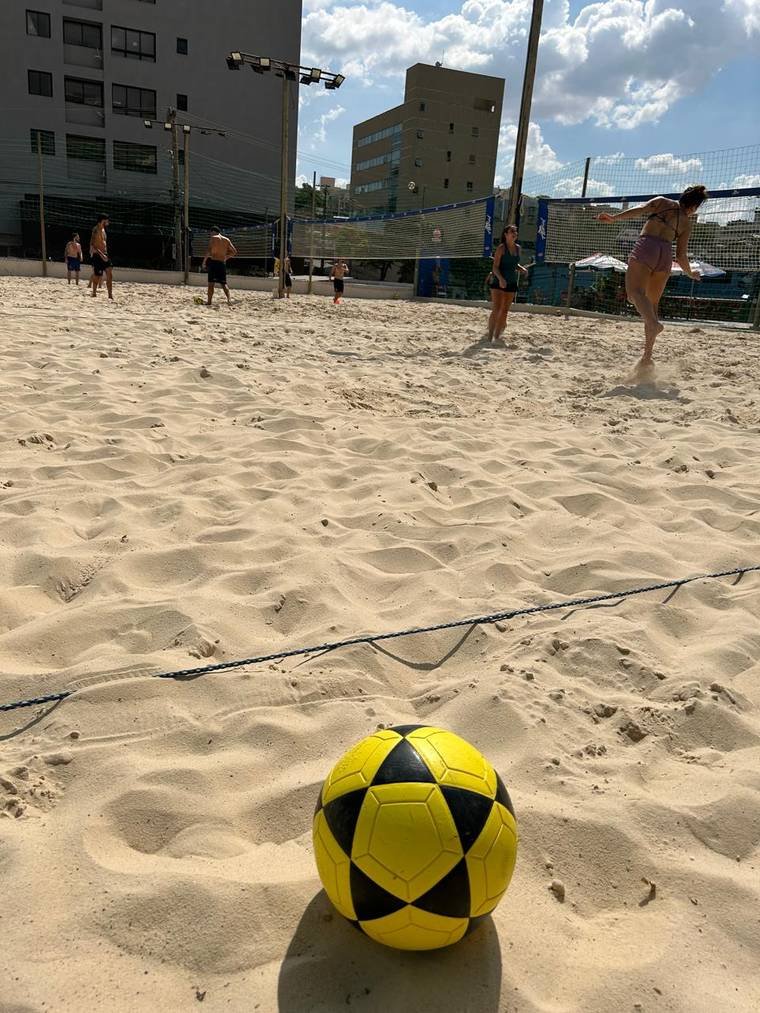 Bola de futevôlei na areia com jogadores ao fundo na quadra da Astral
